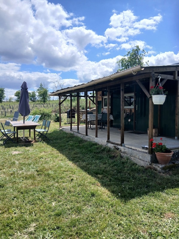 A covered outdoor seating area with a table and chairs, a hanging flower pot, and a view of the surrounding landscape.