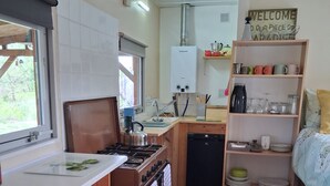 Una cucina compatta con fornello, lavello e un piccolo frigorifero. Una finestra lascia filtrare luce naturale e uno scaffale contiene stoviglie.