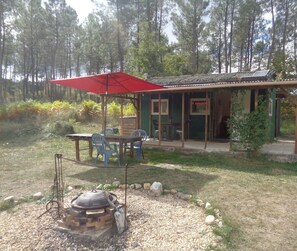 Una cabaña rústica con un espacio de estar cubierto al aire libre, una estufa de leña y vistas a una zona boscosa.