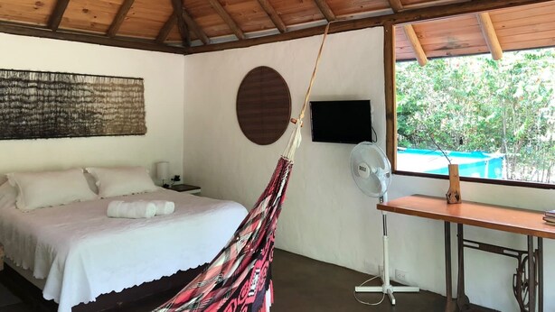 Une chambre avec un lit, un bureau, un ventilateur, une télévision et une fenêtre donnant sur un paysage verdoyant.