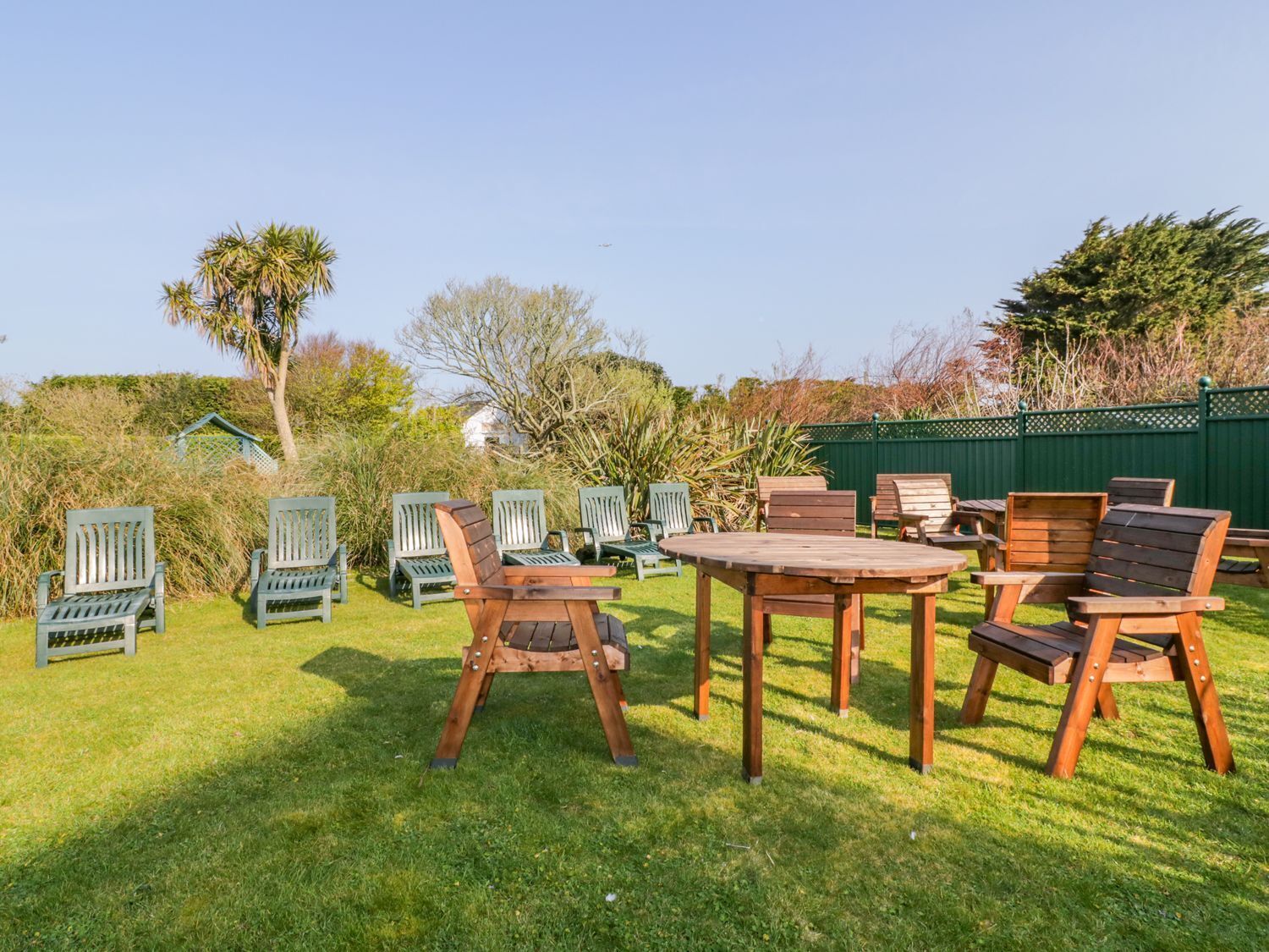 A patio area with wooden chairs and a table, surrounded by greenery and a green fence.