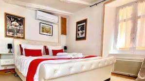 A bedroom with a white bed, red and white bedding, an air conditioning unit, framed pictures on the walls, and a window with curtains. - Studio in the Medieval Center of Sarlat (FRANREAL) (Sarlat la Caneda)