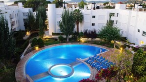 En swimmingpool omgiven av en vackert anlagd trädgård, med utsikt över vita byggnader och ett berg i bakgrunden.