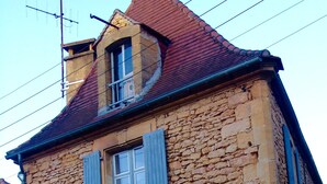 Un edificio de piedra con dos ventanas y contraventanas azules, que presenta un pequeño balcón y una chimenea.