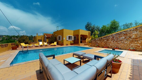 A swimming pool with lounge chairs and a table, surrounded by a yellow building with a stone wall.