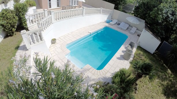A rectangular swimming pool with a white deck, surrounded by a white fence and a small garden area with various plants. - Air-conditioned apartment with large terrace on property with swimming pool (La Roquette-sur-Siagne)