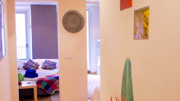 A hallway with wooden flooring, a bed with colorful pillows, a small table, and a wall with framed pictures and a wall hanging. - Modern apartment in Chueca Madrid (Madrid)