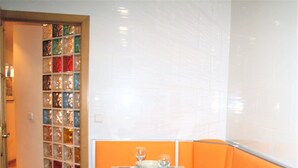 A dining area with a table set for two, featuring a glass partition with colorful glass blocks, orange cushioned seating, and a white tiled wall. - Modern apartment in Chueca Madrid (Madrid)
