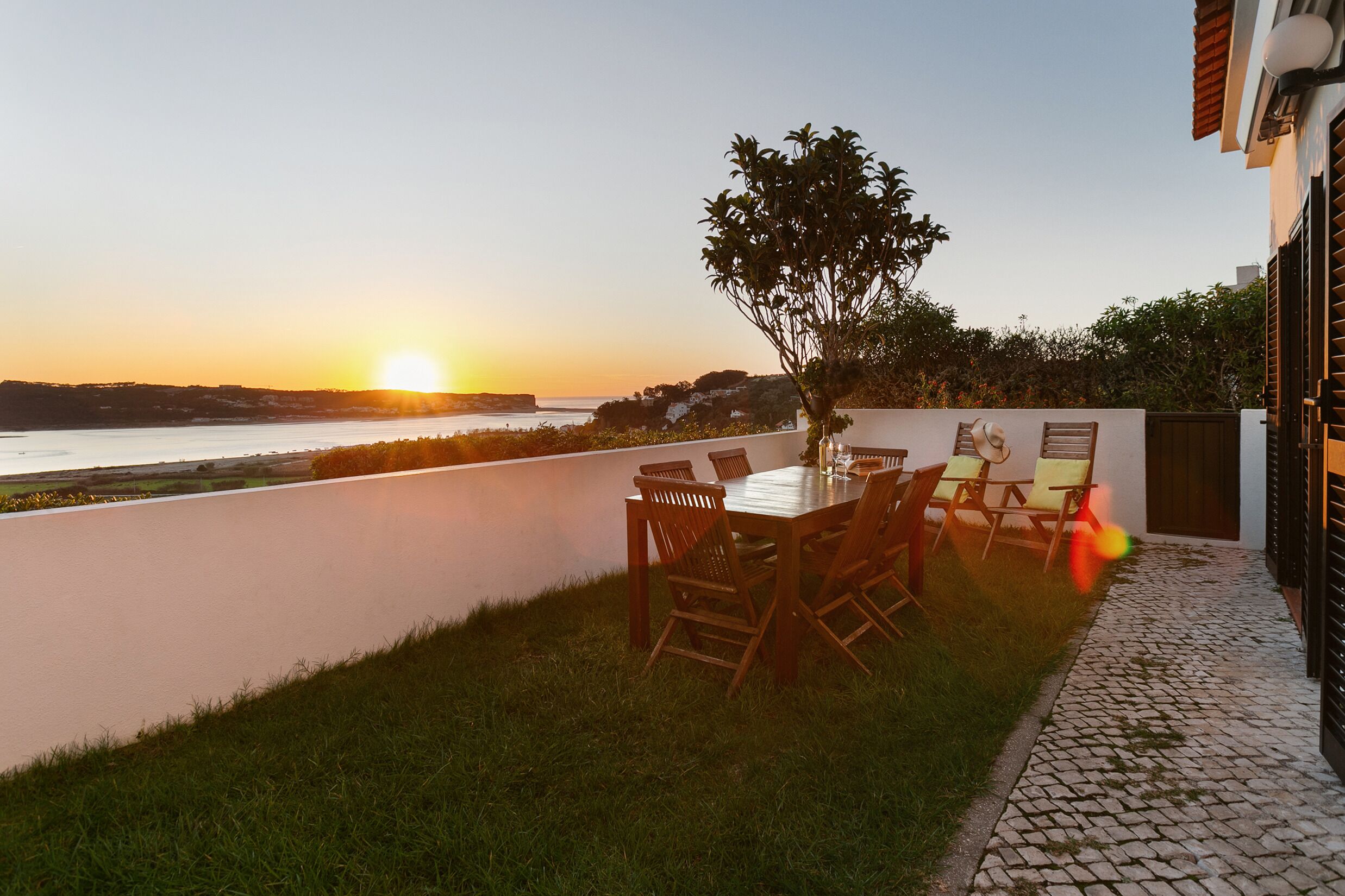 An outdoor patio with a table and chairs, a tree, and a view of the sunset over the water.