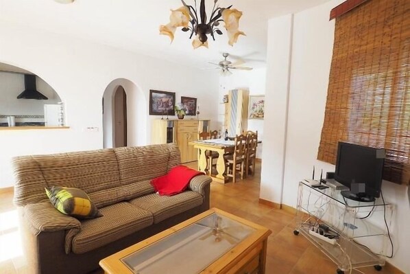 Sala de estar com sofá, mesa de centro, mesa de jantar e cadeiras. Há uma televisão em um suporte e um ventilador de teto.