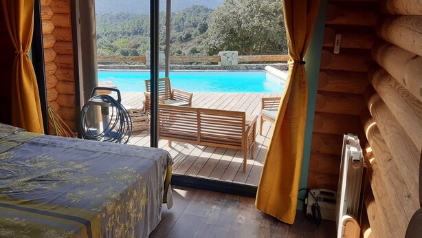 A wooden cabin room with a bed, a table, and chairs near a large window overlooking a pool.