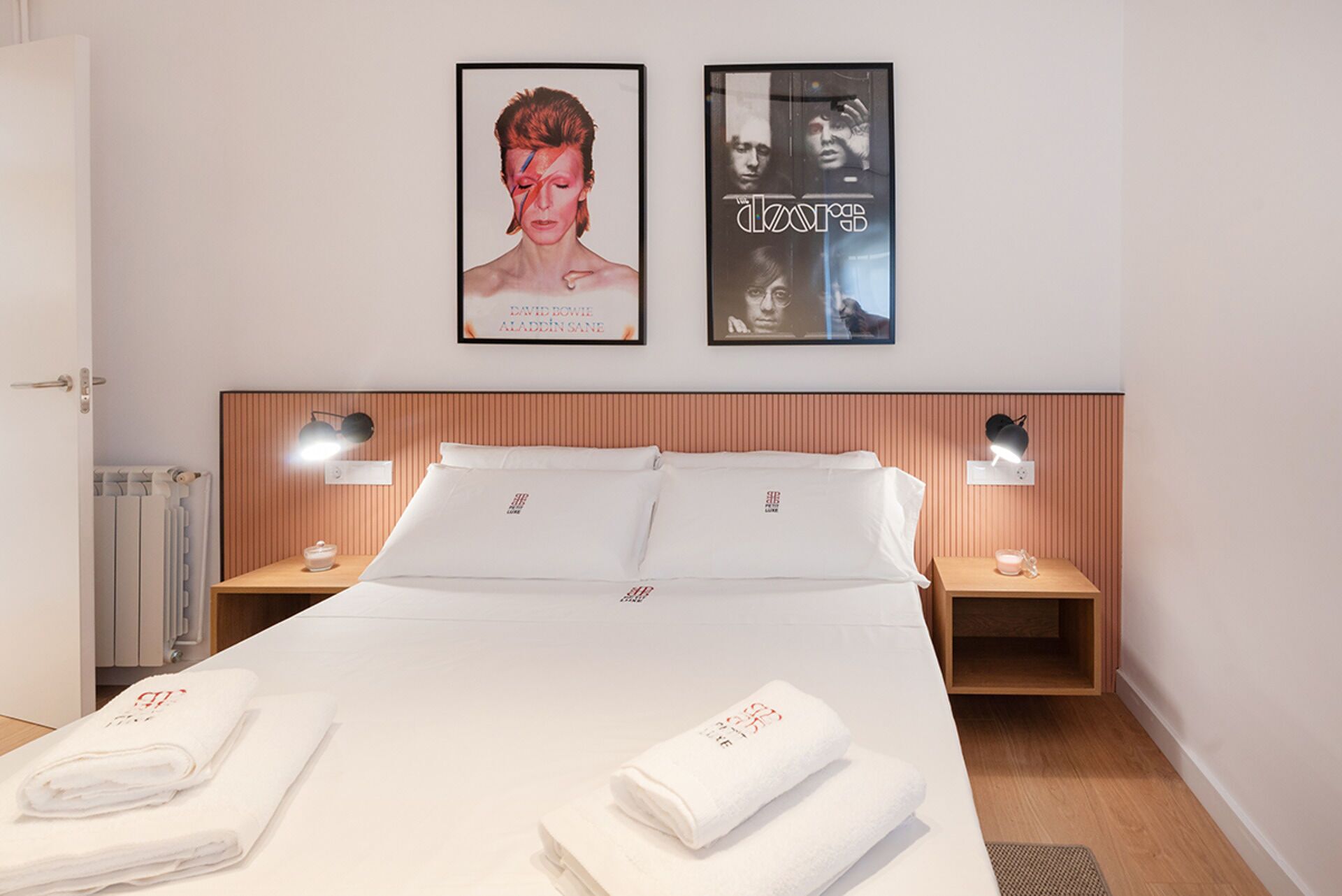 A hotel room with a neatly made bed, two bedside lamps, a poster of a musician, and a radiator.