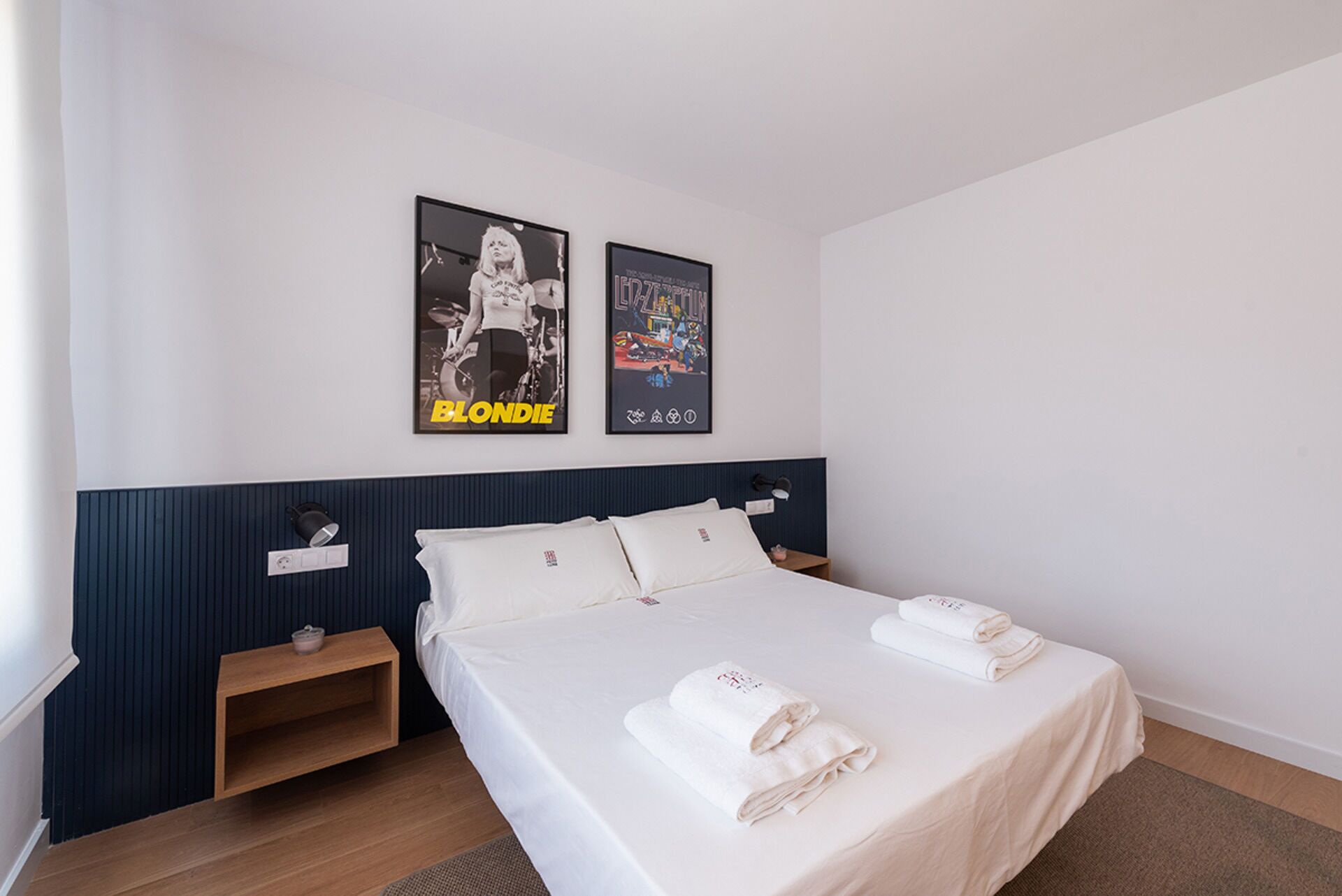 A hotel room with a bed, two pillows, a nightstand, a wall-mounted lamp, and two posters on the wall.
