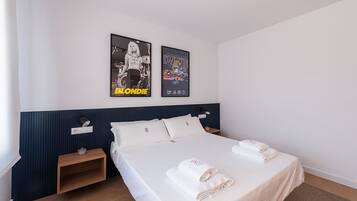A hotel room with a bed, two pillows, a nightstand, a wall-mounted lamp, and two posters on the wall.