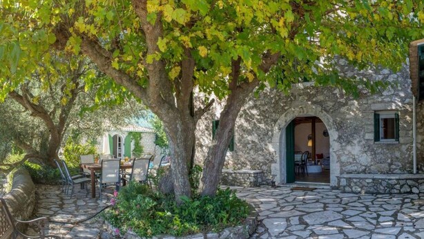 Une cour pavée avec un grand arbre, des sièges extérieurs et un bâtiment rustique doté d’une arche.