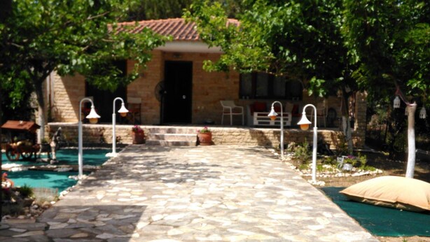 Un chemin de pierres mène à un bâtiment doté d’un toit en tuiles, entouré de verdure et d’une piscine.