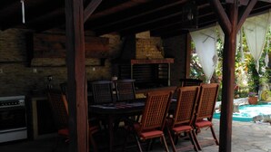 Un patio extérieur couvert, doté d’un sol en pierre, de meubles en bois et d’un espace barbecue.