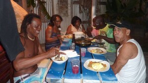 A group of people dining at a wooden table with blue accents. -  "A Village Experience" (Oistins)