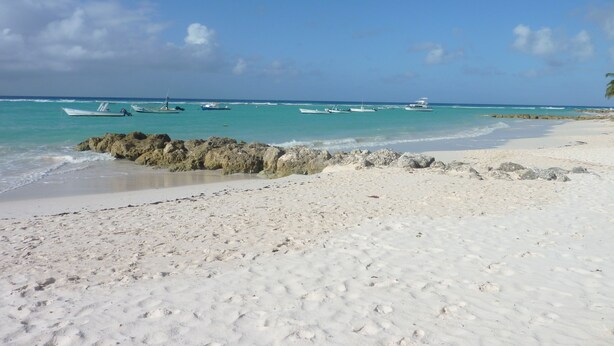 A beach with white sand and turquoise water. There are several boats anchored in the water. -  "A Village Experience" (Oistins)