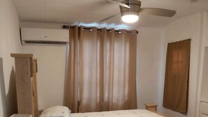 A bedroom with a bed, a ceiling fan, a window with curtains, a bedside table, and a lamp. -  "A Village Experience" (Oistins)