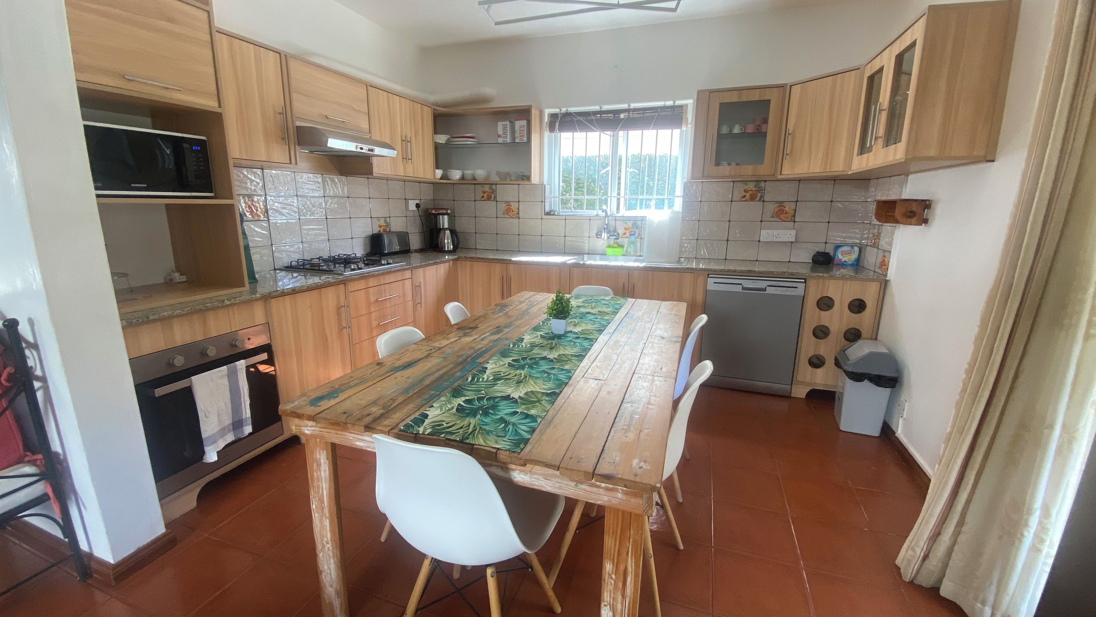 A kitchen with wooden cabinets, a dining table with a leaf-patterned table runner, a dishwasher, and a window with a view.