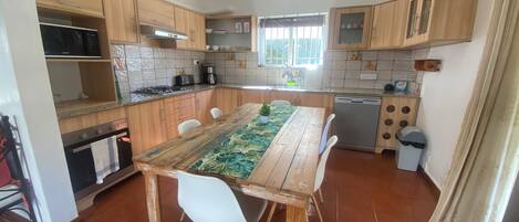 A kitchen with wooden cabinets, a dining table with a leaf-patterned table runner, a dishwasher, and a window with a view.