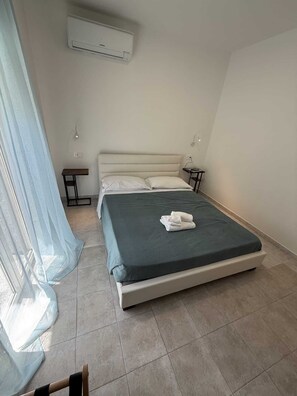 A bedroom with a bed, a bedside table, a nightstand, an air conditioning unit, and a window with sheer curtains. - Family House B Sea Side (letojanni)