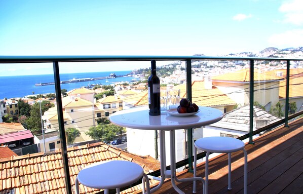 A balcony with a table and two chairs, overlooking a coastal town with buildings and a bridge.