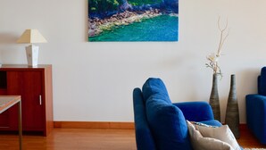 A living room with a blue sofa, a small table with a fruit bowl, a painting of a beach, and a lamp on a wooden cabinet. - Esmee Apartment, Big Terrace with Sea View! (Arco Da Calheta)