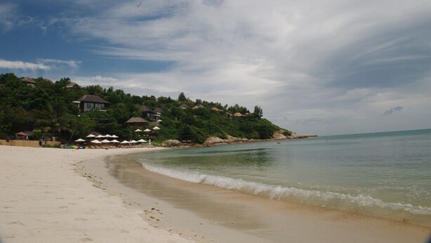 Une plage bordée d’une rangée de parasols et d’une rangée de maisons aux toits de chaume.