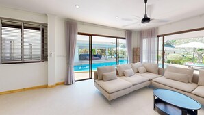 Un salon moderne avec un canapé d’angle beige, une table basse en verre et un ventilateur de plafond. De grandes fenêtres équipées de stores et de rideaux offrent une vue sur un espace piscine extérieur.