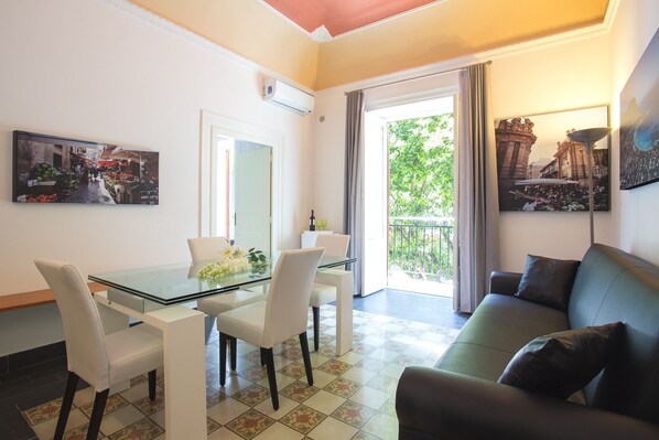 Un salon moderne avec une table à manger en verre, des chaises blanches, un canapé vert et un balcon offrant une vue sur un immeuble.