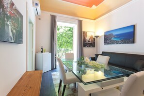 Un salon moderne avec une table à manger en verre, des chaises blanches, un canapé foncé et une vue sur l’extérieur.