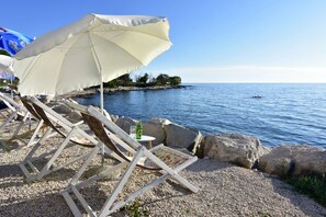 Un entorno frente a la playa con sillones de madera y una sombrilla blanca, con vistas a un mar tranquilo y a una costa rocosa.