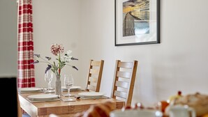 Une table à manger est dressée avec des assiettes, des verres et un vase rempli de fleurs. Un tableau encadré est accroché au mur, et un rideau à carreaux rouges et blancs est visible.