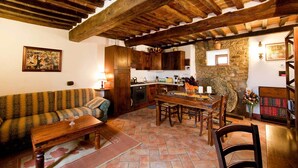 Ein rustikales Interieur mit freiliegenden Holzbalken, einer Steinwand, einer Küche mit Holzschränken, einem Esstisch mit Stühlen und einem gemusterten Sofa.