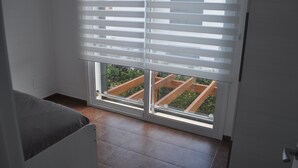 Une chambre avec un lit, une fenêtre avec des stores et une vue sur une terrasse en bois à l’extérieur.