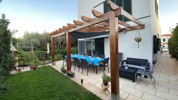 Un espace patio avec une table dressée pour les repas, une structure en forme de pergola et une pelouse bien entretenue.