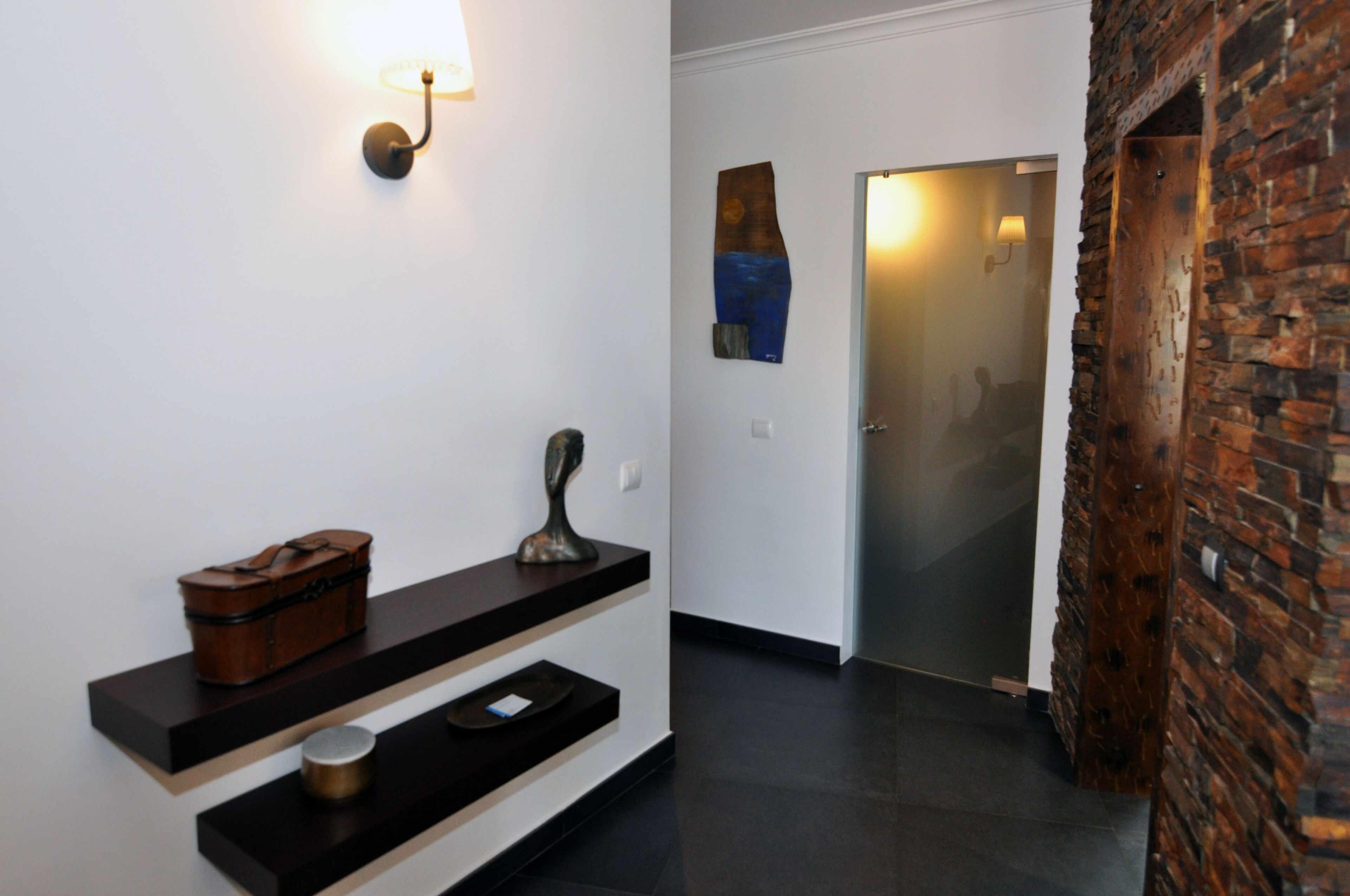 Un couloir avec un banc en bois, un luminaire fixé au mur, une porte avec un panneau de verre et un mur décoratif en briques.