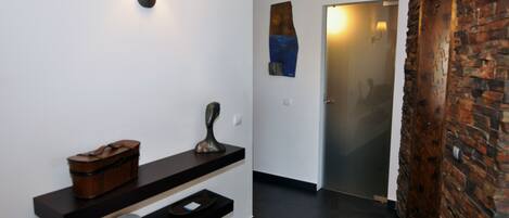 Un couloir avec un banc en bois, un luminaire fixé au mur, une porte avec un panneau de verre et un mur décoratif en briques.