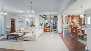 Amplia sala de estar con un sofá grande, una mesa de centro y un comedor con sillas y una mesa. Hay una zona de cocina con armarios de madera y una escalera que conduce a otro nivel.