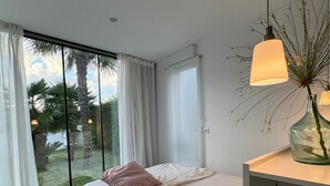 A bedroom with a large bed, a nightstand, a window with curtains, and a view of greenery outside. - House with garden and pool 5 minutes from the beaches (boiro)
