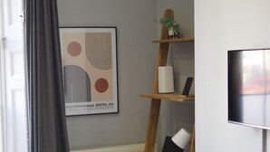A room with a desk, chair, shelves, a radiator, a TV, and a window with curtains.