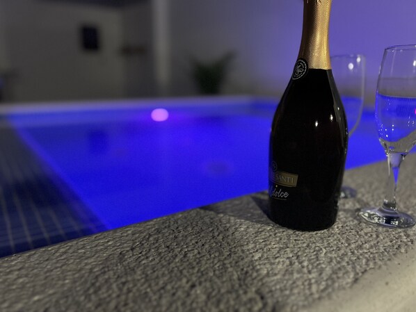 En flaska champagne och ett glas på en stenkant vid en pool med blå belysning.