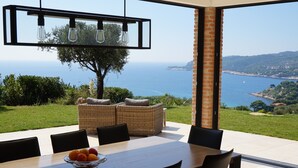 A dining area with a table, chairs, and a bowl of fruit. There is a wicker sofa and a large window with a view of the sea and hills.