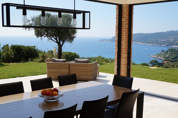 A dining area with a table, chairs, and a bowl of fruit. There is a wicker sofa and a large window with a view of the sea and hills. - Magnificent ancient farm that has been reformed. Exceptional location in Cassis (Cassis)