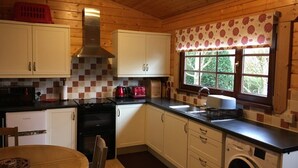 A compact kitchen with wooden cabinets, a built-in oven, a washing machine, and a window with a floral curtain. - Holly Lodge, Cedar Springs, Heacham. 5 Mins Walk to Beach (Heacham)