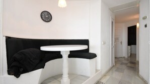 A white room with a round white table, a black bench, a clock on the wall, and a hallway leading to another room.