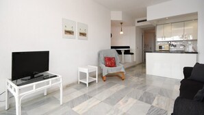 A modern living room with a flat-screen TV on a white stand, a small white side table, a checkered armchair with a red pillow, and a black sofa.
