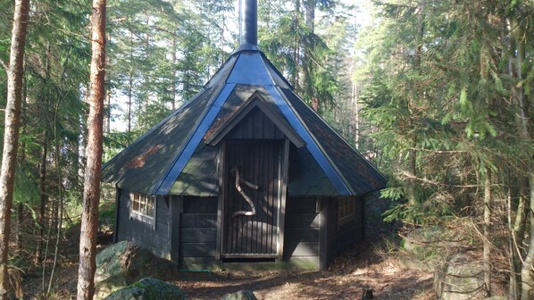 A small cabin with a steeply pitched roof, surrounded by dense forest.